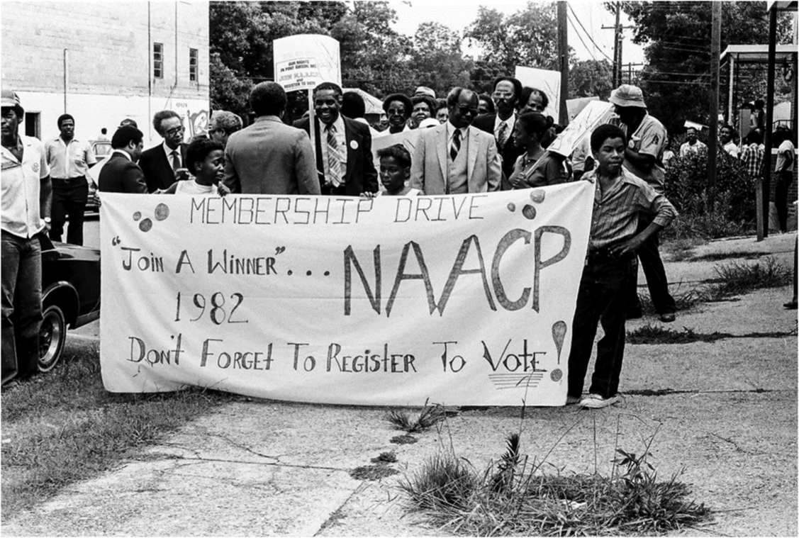 Behind this hand-painted banner, marchers formed up to follow the path of the marches of the 1960s. This re-enactment was joyous and carefree, but marches back then were dangerous. Marchers walked carefully in pairs and had to pass through a gauntlet of hostile and sometimes gun-carrying whites to publicly declare their support for the NAACP demands and their dissatisfaction with the status quo. It was their determination to claim the streets and sidewalks of the town that paved the way for the freedom of access to public places they enjoy today. The children carrying the banner are Angela Weathers, Tawn King, and Winston Moore. Standing left front (in cap and white shirt) is Roderick Devoual; next to him in rear is Percy Thornton, later to be elected a County Supervisor; standing center facing front and carrying a sign is Rev. Eddie Walls, a defendant in the lawsuit, later to be elected a Port Gibson City Alderman; facing him is probably Aaron Henry, president of the state NAACP.
